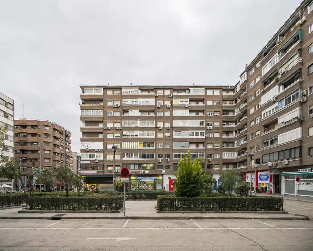 Street-level exterior view of a residential block with storefronts and a central planted median, captured on an overcast day.