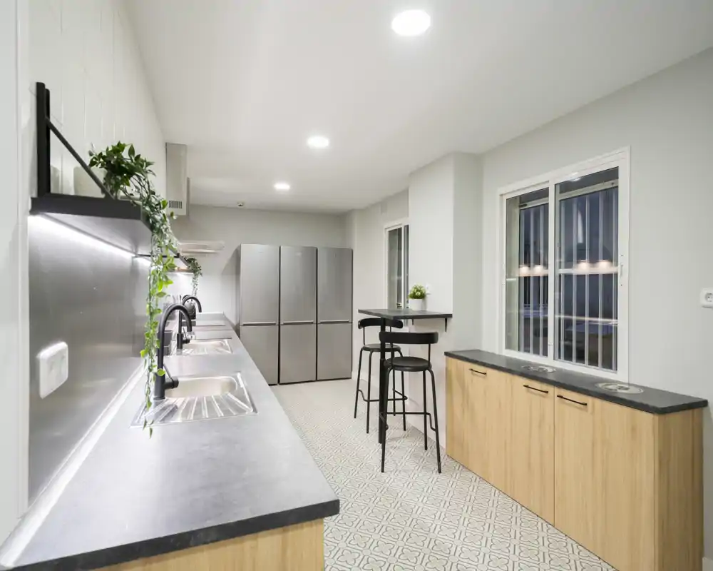 Bright, modern kitchen with long countertops, dual sinks, stainless appliances and a small bar table — sleek wood cabinets and patterned tile flooring.