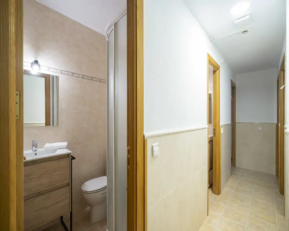 Small bathroom visible from the hallway with a shower cabin, toilet and vanity with mirror. Neutral beige tiles and warm wooden door frames create a clean, functional look.