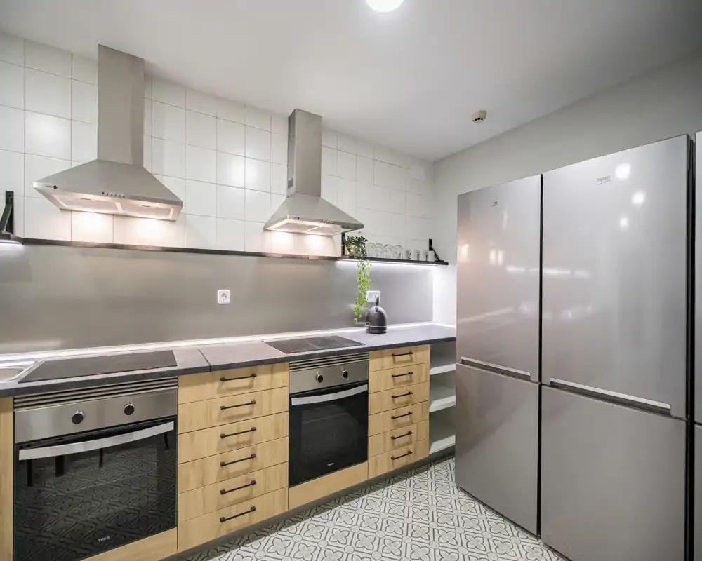 Modern, well-lit kitchen with stainless steel appliances, wooden cabinets, built-in ovens and a large fridge — bright and functional space for cooking.