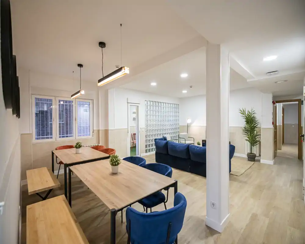 Spacious, modern communal living area with a dining setup and a comfortable blue sofa lounge. Bright, well-lit space with contemporary pendant lights and wood-effect flooring, ideal for shared living.