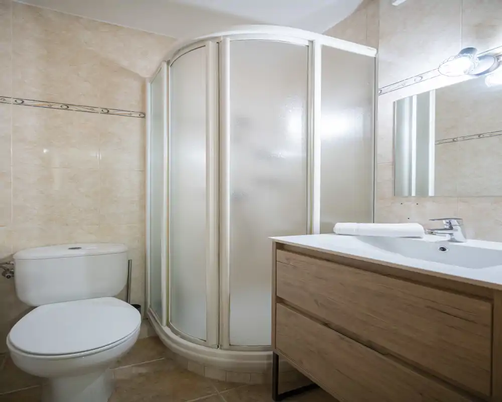 Compact, clean bathroom with a corner shower cabin, toilet and a wooden vanity with sink. Bright, neutral tiles and practical fixtures make it ideal for a rental.