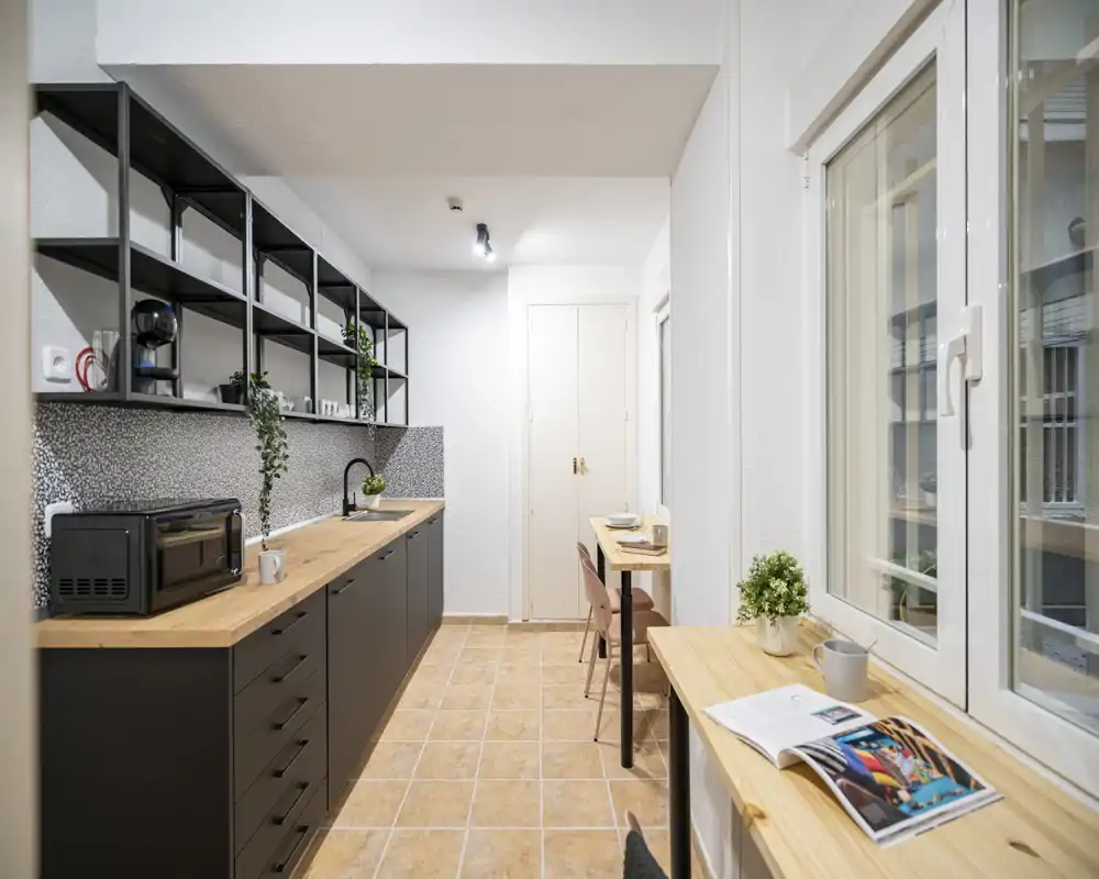 Modern galley kitchen with dark lower cabinets, wooden countertops, built-in sink and open shelving; bright, well-styled space with a small dining/work counter by the window.