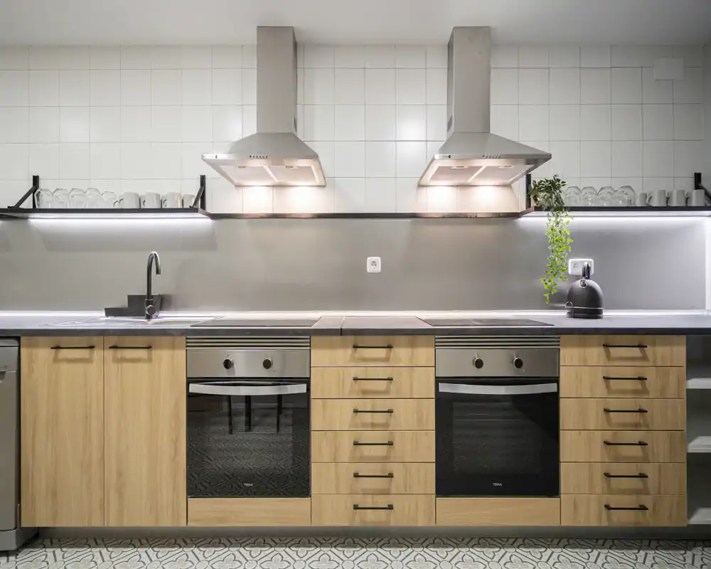 Modern, symmetric kitchen with two built-in ovens, wood-effect cabinets, illuminated extractor hoods and clean tiled backsplash — bright and functional space ideal for cooking.