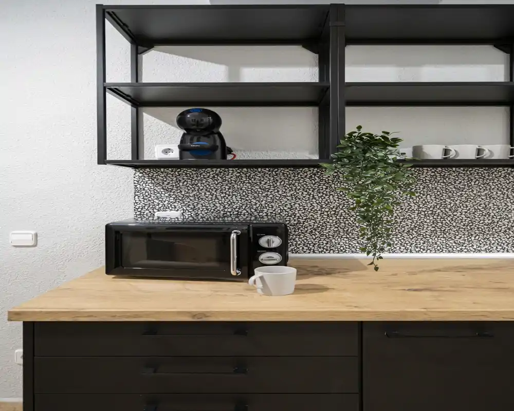 Compact, modern kitchenette featuring a wooden countertop, black microwave, open shelving and a decorative hanging plant — neat and well-styled for short-stay rentals.