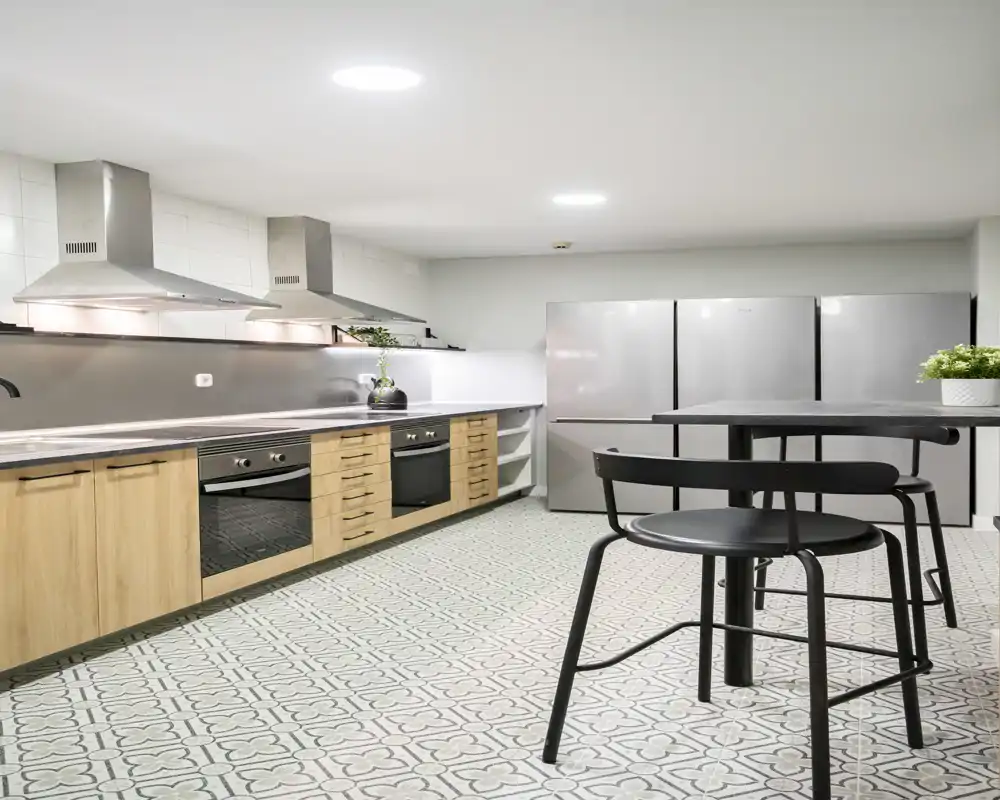 Modern open kitchen with wooden lower cabinets, stainless steel appliances and a small bar table; patterned floor tiles and bright overhead lighting create a clean, functional cooking space.