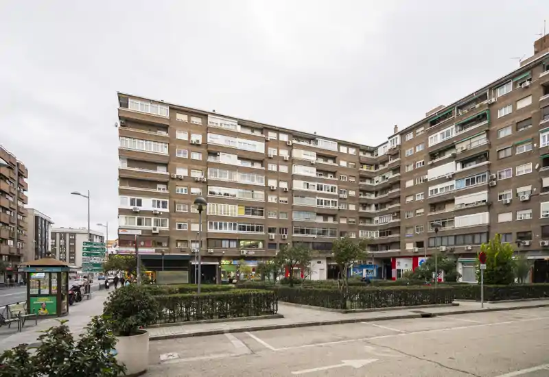 Vue extérieure urbaine montrant la façade d'un immeuble résidentiel, des commerces au rez-de-chaussée et une petite zone plantée sous un ciel couvert.