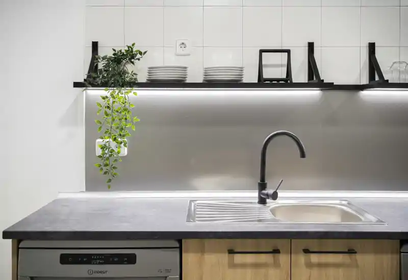 Coin cuisine moderne avec évier en acier inoxydable, robinet noir mat, éclairage sous étagère et petite plante suspendue, présenté dans un style minimaliste et épuré.