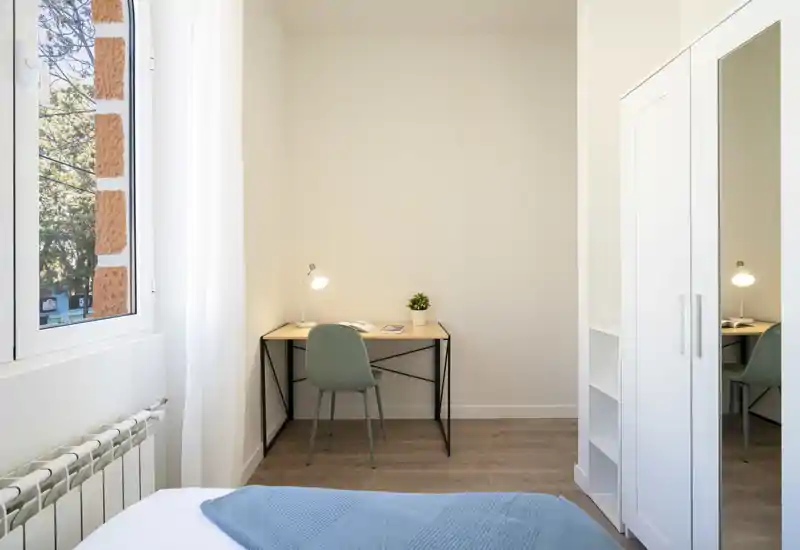 Rincón de dormitorio luminoso y minimalista con el borde de la cama visible, un escritorio pequeño con silla, armario con espejo y una ventana que aporta luz natural.