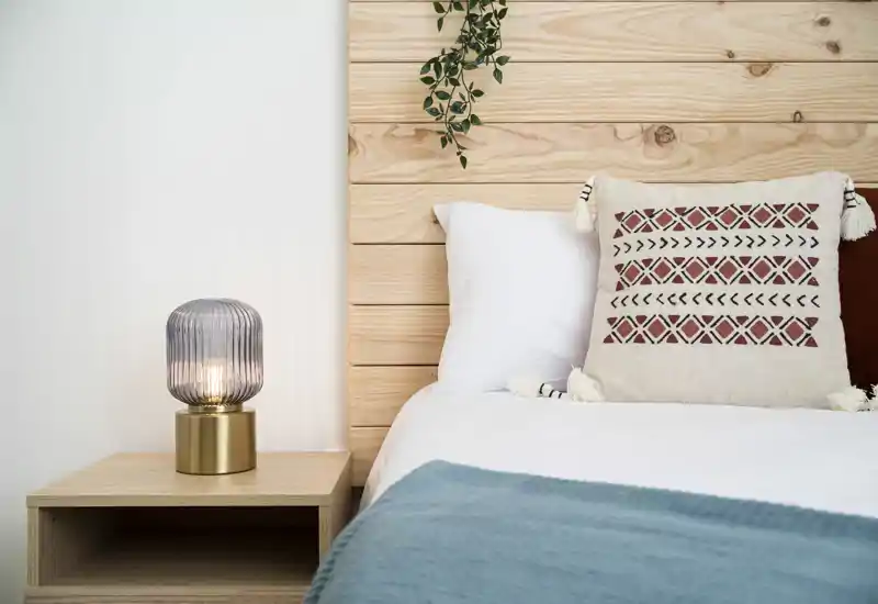 Bright, styled bedroom corner with a wooden headboard, patterned cushion and bedside lamp — a cozy, modern sleeping space.
