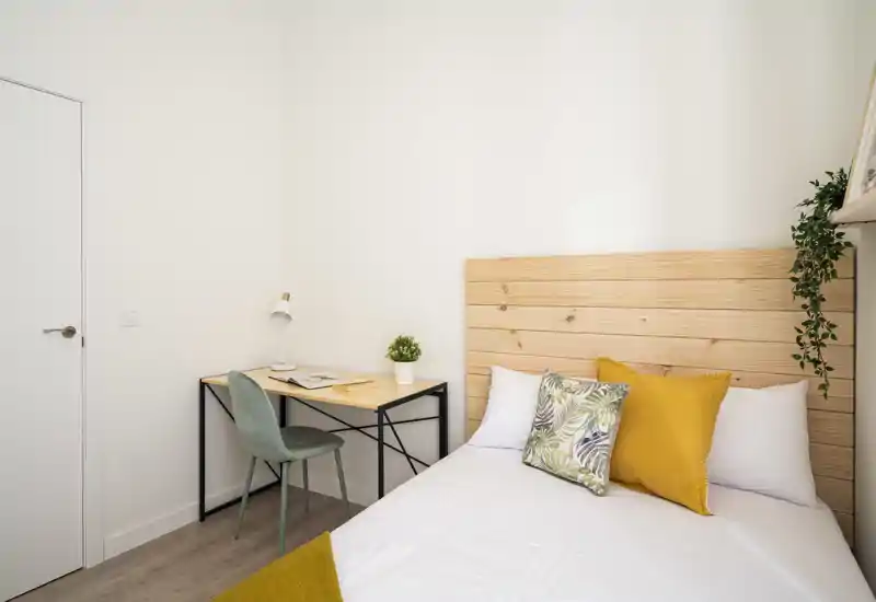 Bright, minimal bedroom with a wooden headboard, white linens and decorative cushions. A small desk and chair sit beside the bed, and a potted plant adds a touch of greenery.