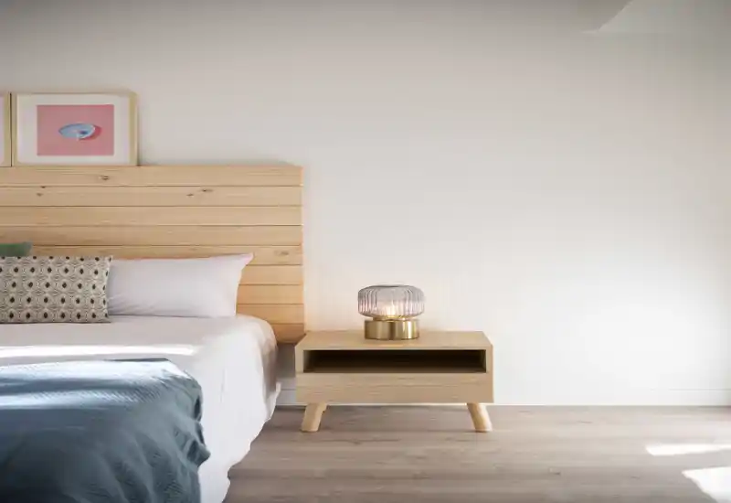 Camera da letto luminosa e minimal con testiera in legno, comodino e una lampada dal design morbido. Il letto è ben rifatto con plaid e cuscini decorativi, la stanza è illuminata da luce naturale.