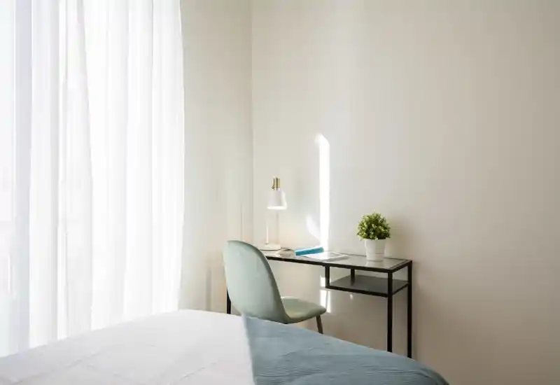 Rincón de dormitorio luminoso y minimalista con cama bien hecha, pequeña mesa de trabajo, lámpara y planta junto a una gran ventana con cortina traslúcida — ambiente relajante.
