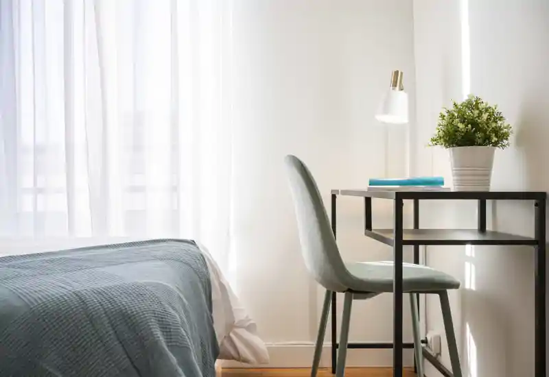 Rincón de dormitorio luminoso y minimalista con cama bien hecha y un pequeño escritorio junto a la ventana — ideal para descansar y trabajar desde casa.