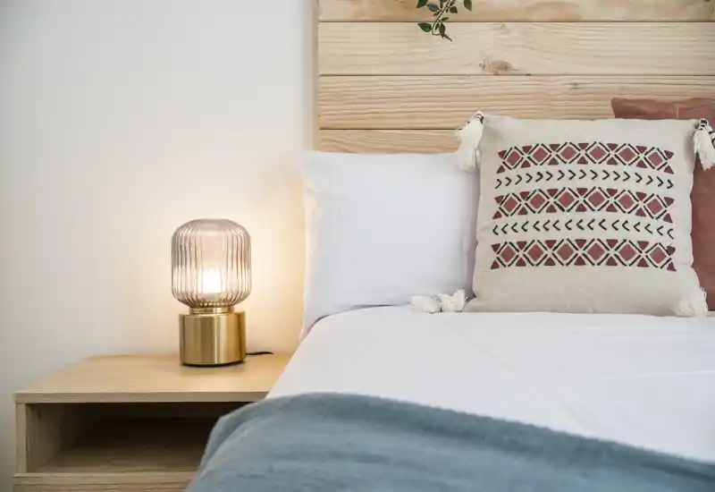 Rincón de dormitorio luminoso y decorado con cama bien hecha, cojines estampados y una cálida lámpara de mesita de noche sobre una mesita de madera.