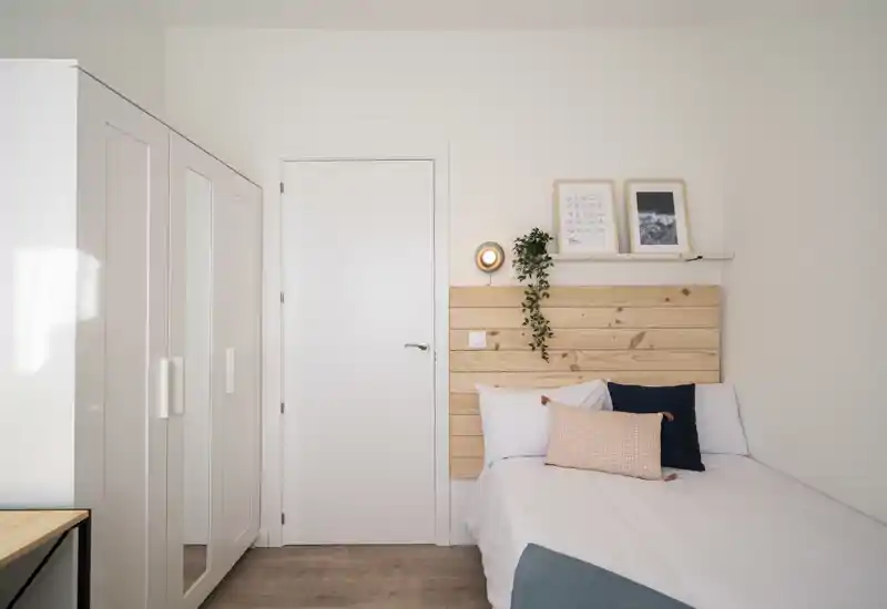 Bright, minimal bedroom with a white wardrobe, wooden headboard and neatly made single bed dressed in white linens and accent cushions.