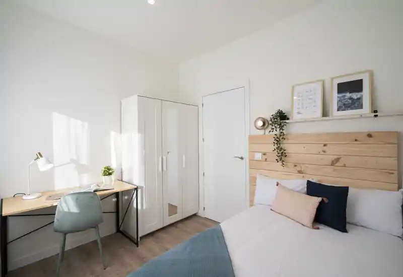 Bright, modern bedroom with a wooden headboard, neatly made bed with decorative cushions and a compact workspace by the window — ideal for a cozy rental room.