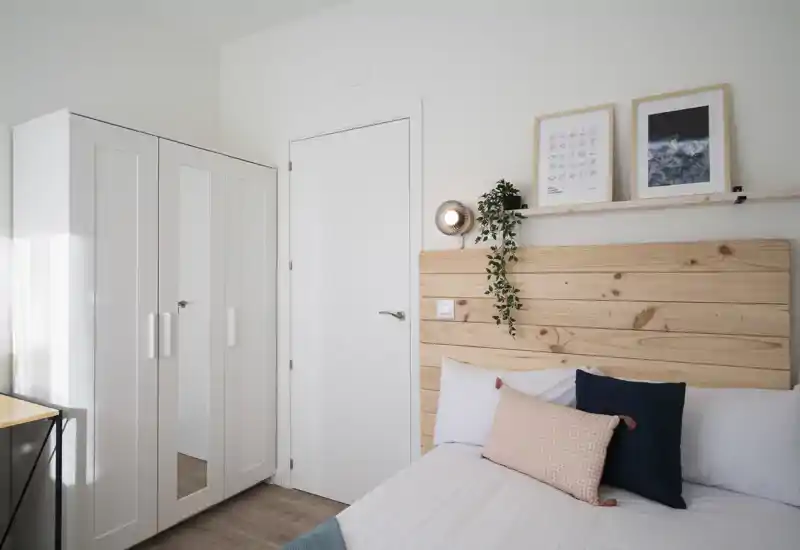 Bright, minimalist bedroom with a wooden headboard, decorative pillows and a white wardrobe — cozy, well-lit and neatly styled.