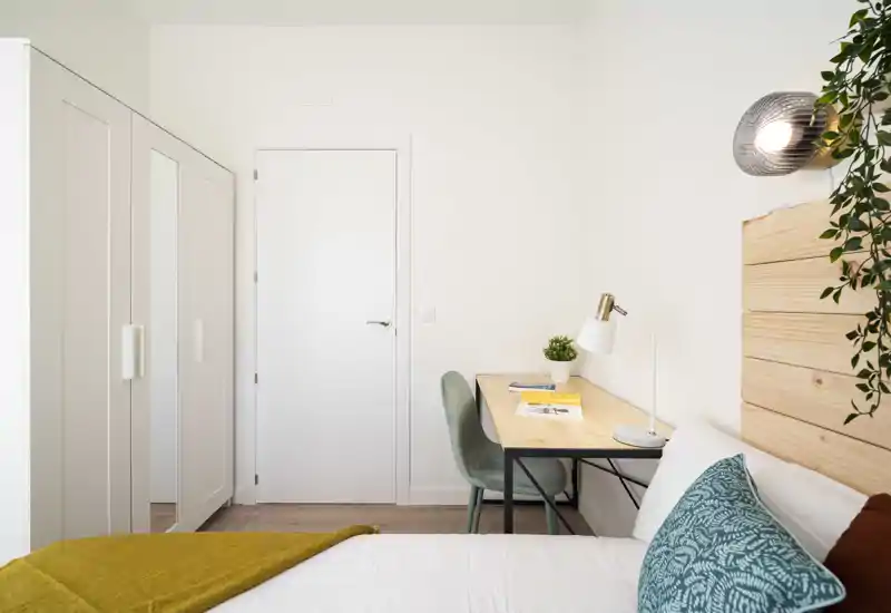 Chambre lumineuse et moderne avec tête de lit en bois, coussins verts et un bureau soigné à côté du lit — idéale pour une location compacte et élégante.