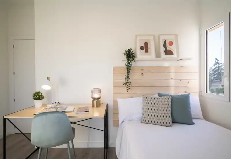 Bright, minimal bedroom with a wooden headboard, decorative pillows and a small work desk by the bed. Natural light from the window and soft neutral tones create a calm, welcoming atmosphere.