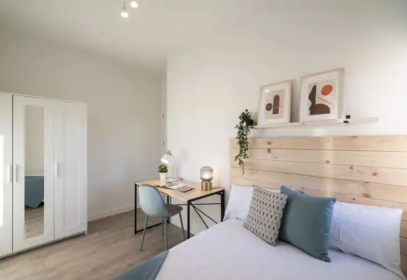 Bright, modern bedroom with a wooden headboard, neatly made double bed, decorative cushions and a small desk by the wall — ideal for a comfortable stay or remote work.