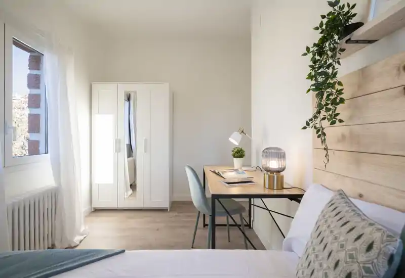 Bright, well-staged bedroom with a wooden headboard, bedside lamp and a small desk by the window — ideal for a cozy urban stay.