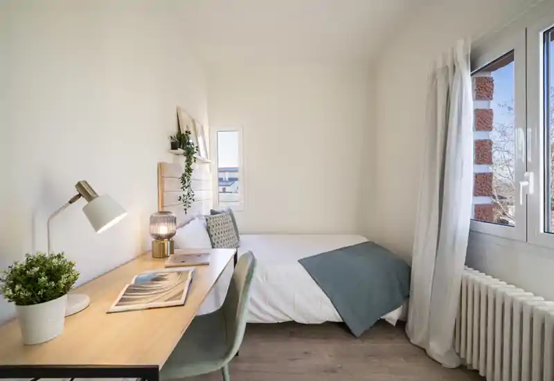 Small bright bedroom with a comfortable single bed, wooden headboard and a tidy desk by the window — ideal for a student or short stay. Natural light fills the room and tasteful decor creates a calm, welcoming atmosphere.
