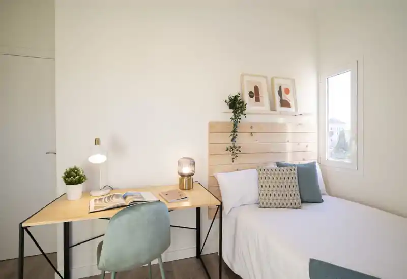 Bright, minimalist bedroom with a wooden headboard, white linens and decorative cushions. A compact desk with a lamp and chair sits beside the bed, ideal for work or study.