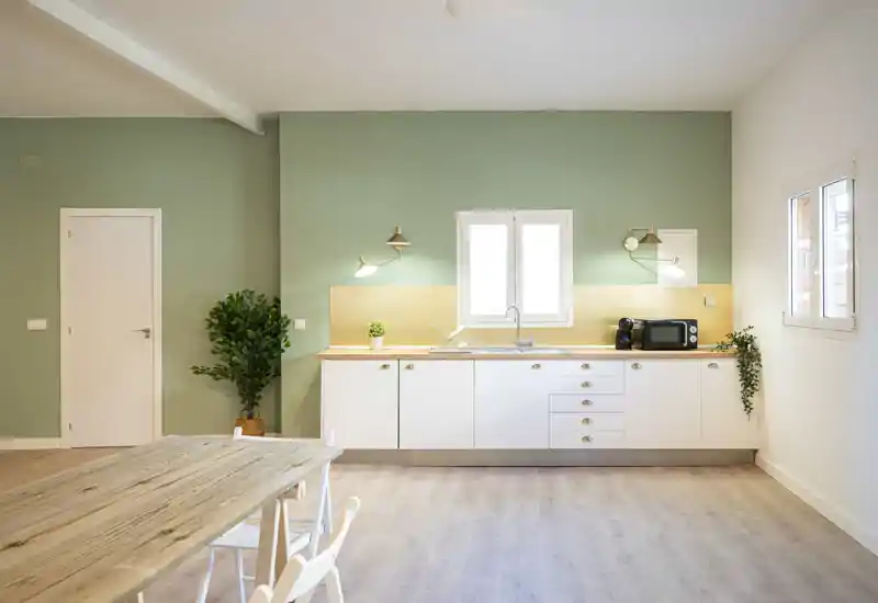Cocina moderna y luminosa con encimera de madera, armarios blancos, fregadero bajo la ventana central y pared verde suave — zona de cocina minimalista y acogedora.
