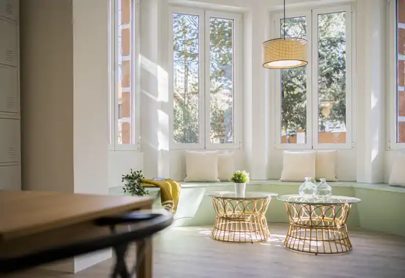 Zona de estar luminosa y acogedora con banco junto a la ventana, cojines y luz natural. Mesas de centro de ratán y tonos neutros crean un ambiente cálido y relajante.