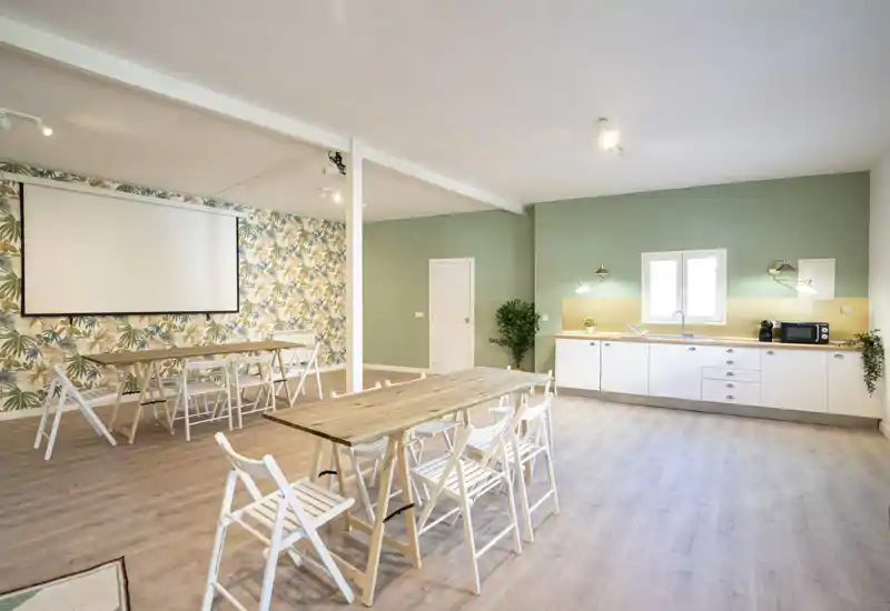 Amplia cocina de concepto abierto con zona de comedor, suelos de madera clara, una larga encimera con armarios blancos y varias mesas de madera con sillas plegables — ideal para comidas en grupo o eventos.