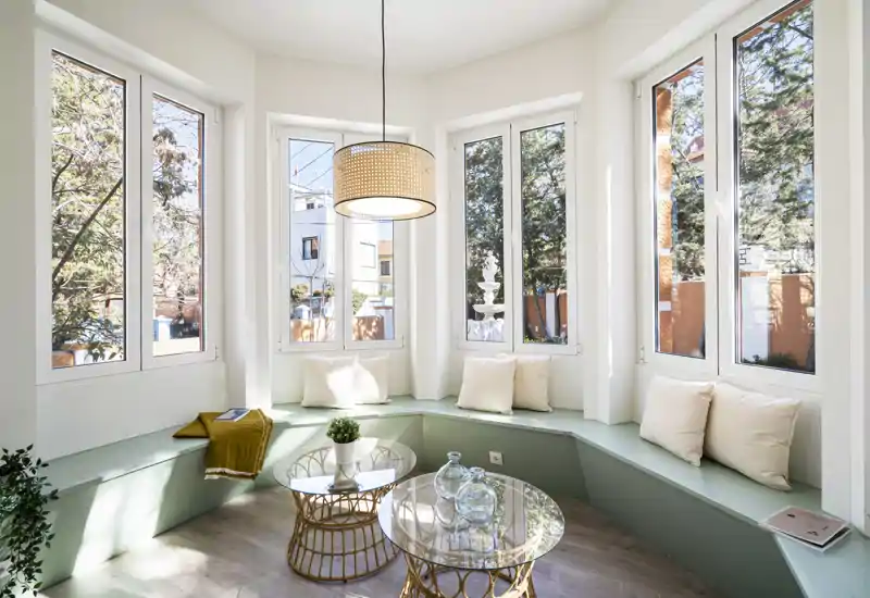 Rincón de salón luminoso con asiento integrado bajo amplias ventanas, cojines y mesas con sobre de cristal — ideal para relajarse o leer con luz natural.