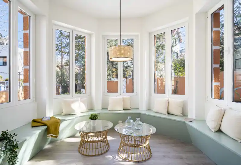 Acogedor rincón con banco envolvente, cojines, mesas de cristal y grandes ventanas que inundan el espacio de luz natural — perfecto para relajarse o leer.