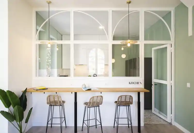 Cocina moderna y luminosa con barra de madera, tres taburetes de ratán y lámparas colgantes; amplios ventanales interiores que crean sensación de amplitud.