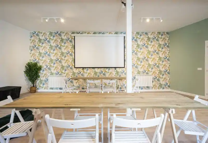 Zona de comedor luminosa y decorada con una larga mesa de madera, sillas plegables blancas y papel pintado botánico — ideal para comidas en grupo o reuniones.