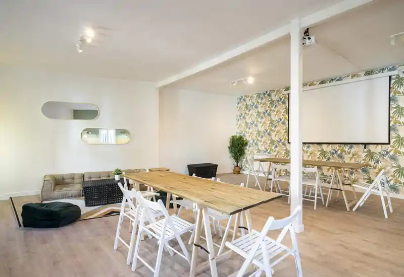 Área común luminosa y con estilo con una larga mesa de madera, sillas plegables, zona de descanso y papel pintado decorativo — ideal para reuniones o encuentros sociales.