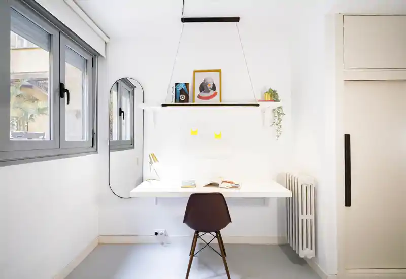 Rincón de oficina en casa luminoso y minimalista con escritorio blanco de pared, silla de diseño, estante y un espejo junto a una gran ventana que aporta luz natural.