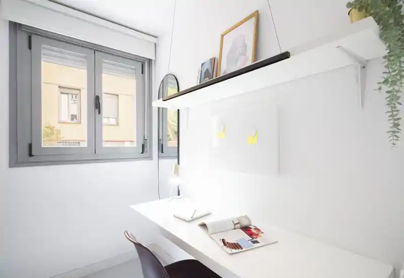 Espacio de trabajo minimalista y luminoso junto a una ventana grande, con escritorio blanco, lámpara y estantería — ideal para estudio o teletrabajo.
