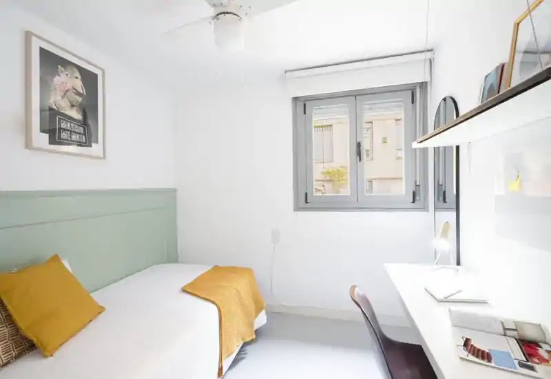 Bright, compact bedroom with a single bed dressed in warm textiles, a desk by the window and clean minimalist decor — ideal for a student or single occupant.