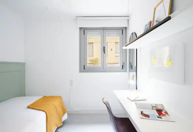 Bright, minimalist bedroom with a single bed, a neat desk and a window providing natural light. Clean lines and a warm throw create a calm, functional space ideal for a solo renter.