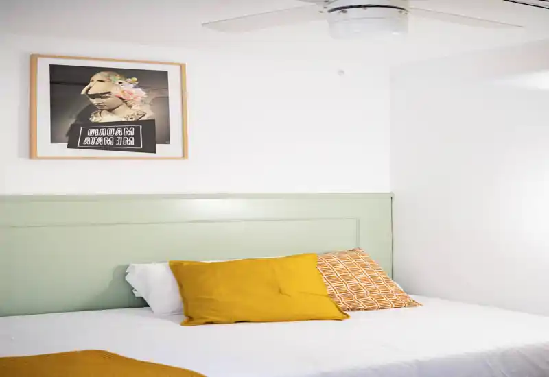 Bright, styled bedroom with a large bed dressed in white linens and warm mustard cushions, framed artwork above a pale green headboard and a ceiling fan — cozy and modern.
