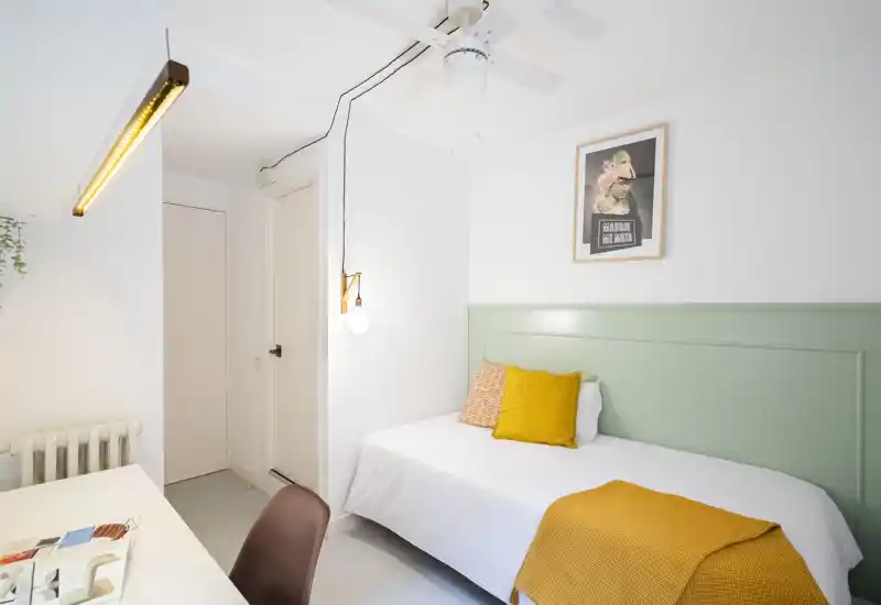 Compact, well-lit bedroom with a single bed, soft green headboard and warm mustard accents. Clean, minimal styling ideal for a short-term rental or guest room.