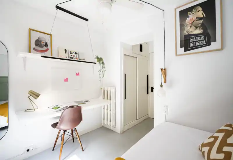 Angolo camera compatto e di design con scrivania a muro, lampada decorativa e letto parzialmente visibile con cuscino decorato. Pareti bianche luminose e quadri conferiscono un'atmosfera moderna e accogliente, perfetta per affitti brevi.