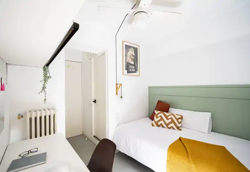 Camera da letto luminosa e minimale con letto singolo, testiera verde, cuscini decorativi e coperta senape. Pareti bianche pulite, angolo scrivania e luce naturale la rendono perfetta per affitti brevi.