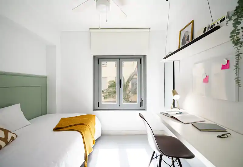 Camera da letto luminosa e minimal con letto comodo, coperte decorative e una scrivania ordinata vicino alla finestra. Luce naturale e pareti bianche donano un’atmosfera ariosa e moderna, perfetta per un annuncio.
