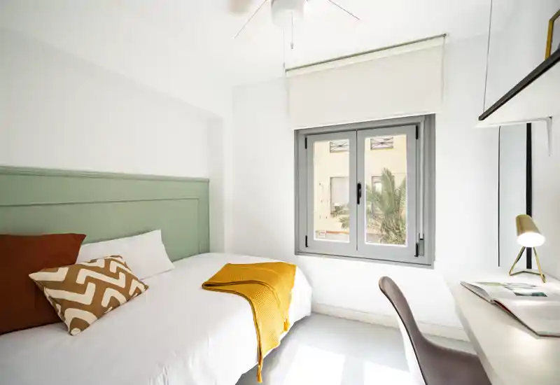 Camera singola moderna e luminosa con letto ordinato, cuscini decorativi e una calda coperta color senape; la luce naturale entra dalla finestra accanto a una scrivania compatta.