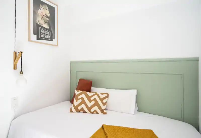 Camera da letto luminosa con testiera verde menta, cuscini decorativi e plaid giallo senape — ambiente curato e accogliente, ideale per affitti brevi.