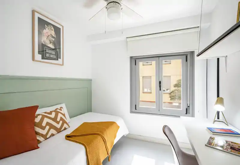 Dormitorio compacto y luminoso con cama individual, cabecero, cojines decorativos y una manta cálida. La ventana proporciona luz natural abundante y hay un escritorio pequeño para trabajar.