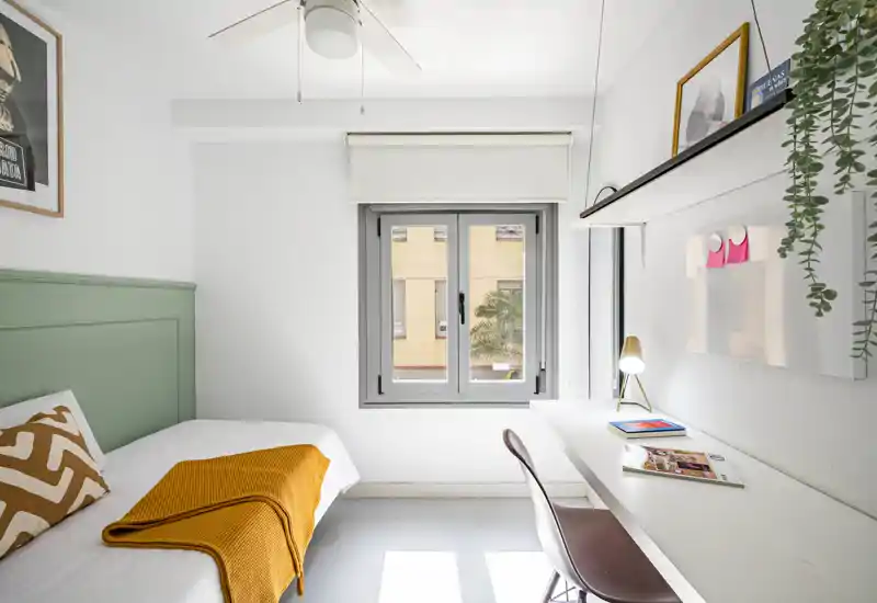 Dormitorio luminoso y minimalista con una cama individual, una manta mostaza acogedora y un área de trabajo junto a la ventana — ideal para estancias individuales o estudiantes.