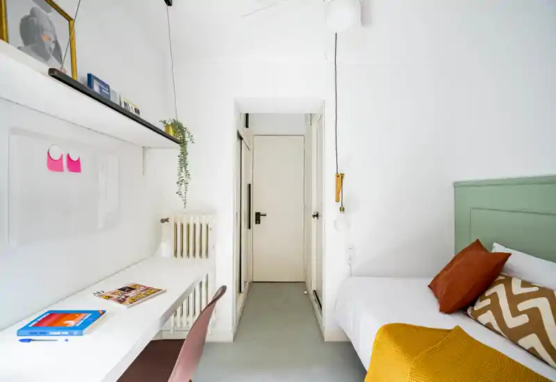 Dormitorio luminoso y compacto con cama individual, cojines cálidos y una manta amarilla, además de un escritorio largo junto a la pared — ideal para estudiante o estancias cortas.